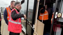 A man in a high-vis vest wearing sight loss simulating glasses is guided onto a tram by another man in a high-vis vest.