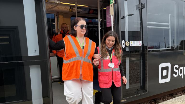 a woman wearing a high-vis vest guides another woman wearing sight-loss simulating glasses off a tram