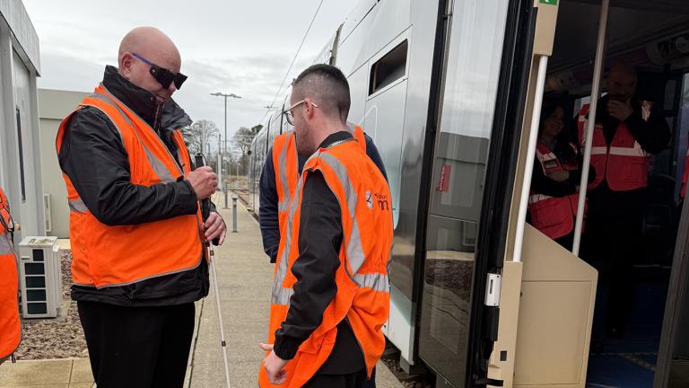 A man wearing a high-vis vest holds a white cane and wears sight-loss simulating glasses, he is speaking to two men wearing high-vis vests outside a tram