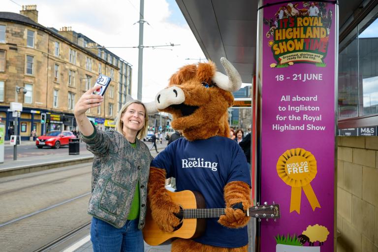 The Royal Highland Show mascot, a Highland Cow, plays guitar on a tram stop advertising the show, a woman takes a selfie with him next to him.