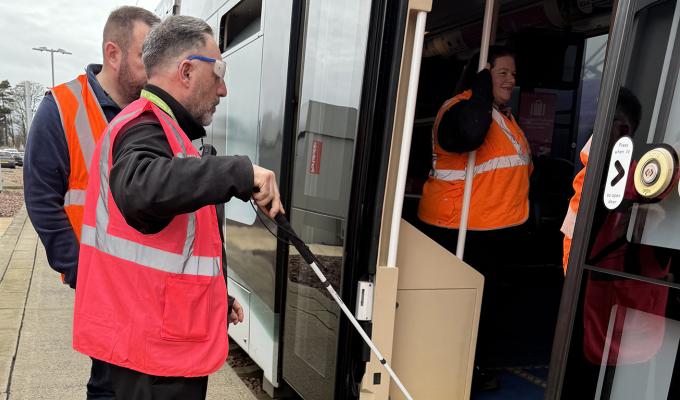 A man in a high-vis vest wearing sight loss simulating glasses is guided onto a tram by another man in a high-vis vest.